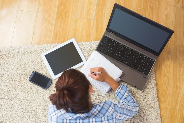 Brunette young student using her tablet and notebook for doing homework lying in the living room.jpeg